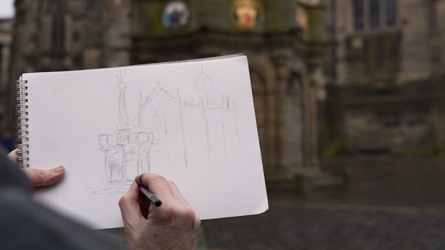 A sketchbook held in front of the Edinburgh Mercat Cross, a hand sketching the scene in pencil.