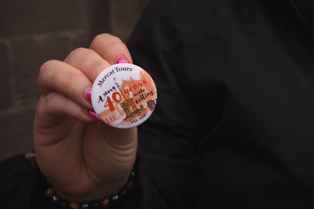 A hand holding up a button with an illustration of the Mercat Cross and text that reads 'Mercat Tours; A story 40 years in the telling; est. 1985’.