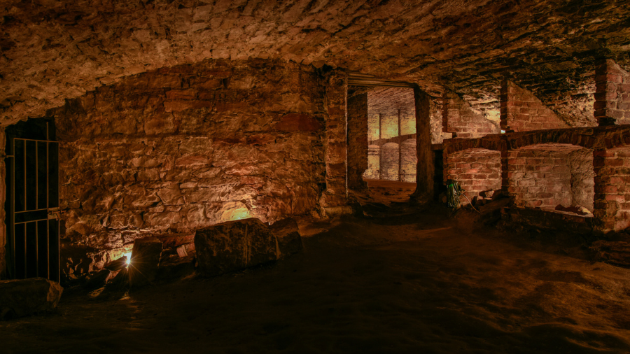 An arched room of the Edinburgh vaults with storage shelves along the right wall and an iron gate in the left corner.