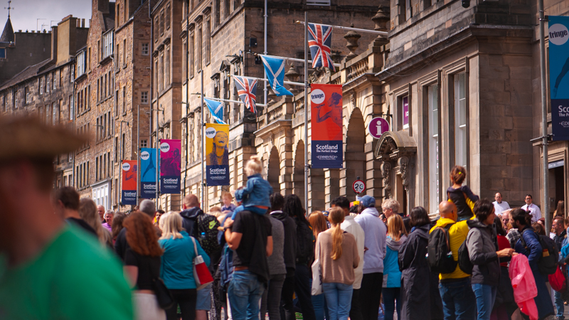 A large group of people gather on the Royal Mile for a street performance, Fringe Festival posters overhead. 
