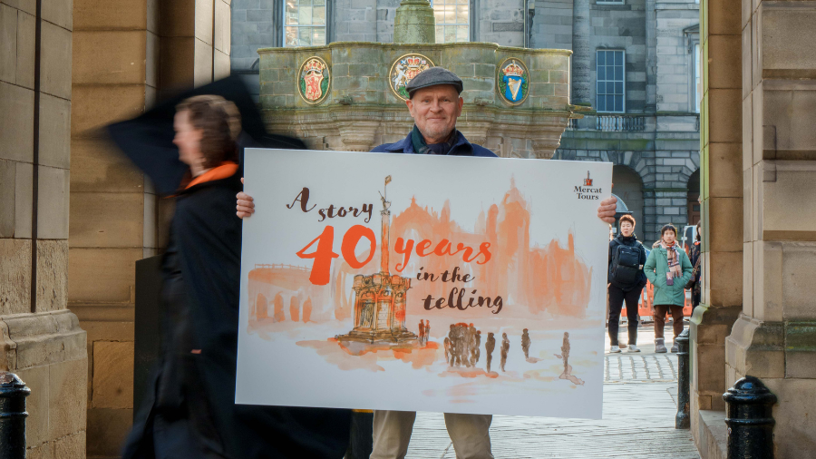 Alasdair MacLauchlan and a Mercat Storyteller stand in front of the Mercat Cross, holding a large watercolour illustration of the Mercat Cross and St Giles Cathedral with script text reading 'A story 40 years in the telling'.