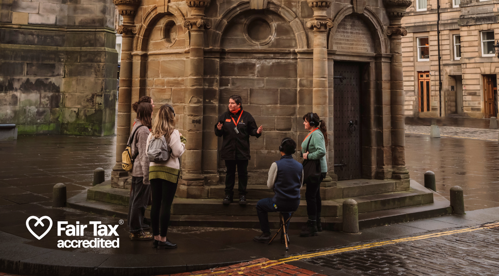 A Mercat Storyteller begins a tour with a group of 5 people at the Mercat Cross, a white 'Fair Tax accredited' logo in the bottom left corner.