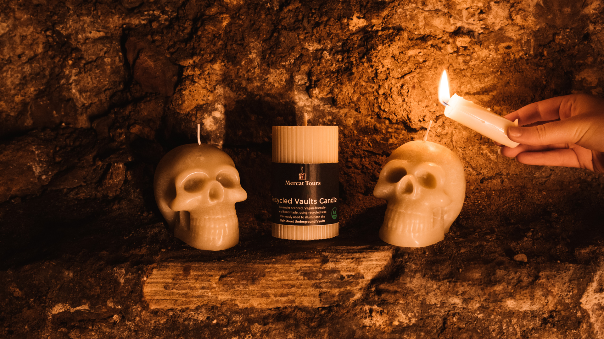 Two candles shaped like skulls and a pillar candle labelled ‘Recycled Vaults Candle’ sit on a ledge in the Edinburgh underground vaults, being lit by another candle. 