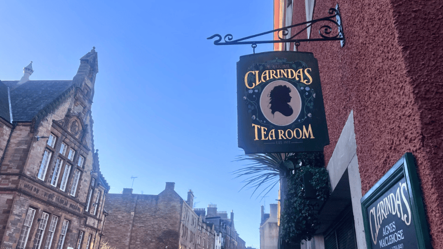 A sign that reads ‘Clarinda’s Tea Room’ with the silhouette of a woman on it against a blue sky and the Edinburgh Royal Mile.